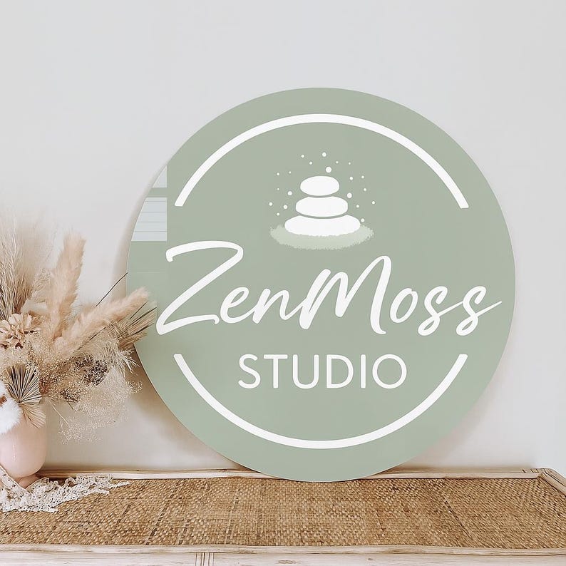 May include: A round, sage green sign with the text "ZenMoss STUDIO" in white script. The sign features a white graphic of stacked stones and is displayed near dried flowers in a vase.