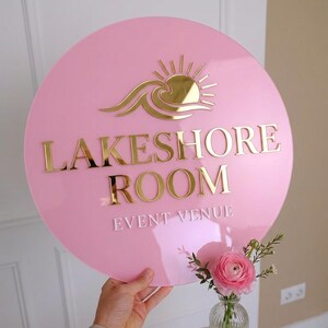 May include: A circular pink acrylic sign featuring a gold mirrored sun and wave icon at the top. The text LAKESHORE ROOM is in gold mirrored lettering, and the words EVENT VENUE are in white lettering below. The sign has a smooth, glossy finish.