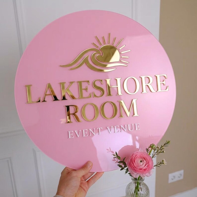 May include: A circular pink acrylic sign featuring a gold mirrored sun and wave icon at the top. The text LAKESHORE ROOM is in gold mirrored lettering, and the words EVENT VENUE are in white lettering below. The sign has a smooth, glossy finish.