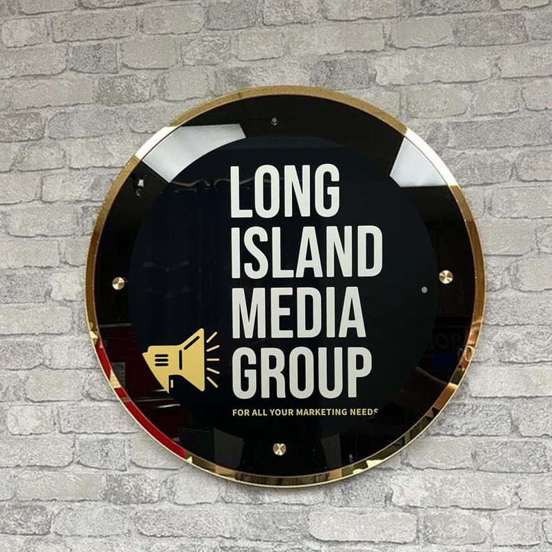 May include: A gold-rimmed round sign with a black background and white text that reads "Long Island Media Group" with a small speaker icon and the text "For all your marketing needs".