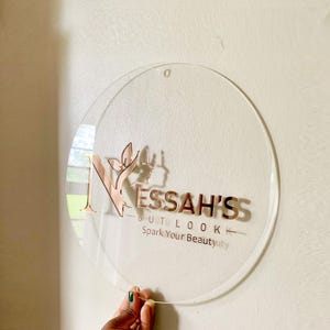May include: A round, clear acrylic sign with a rose gold logo that reads "NESSAH'S OUTLOOK Spark Your Beauty".