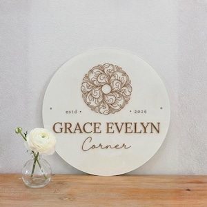 May include: A circular white sign featuring a brown floral mandala design at the top, the text "estd 2026" in the center, and the words "GRACE EVELYN Corner" printed below in brown. A small clear glass vase holds a single white ranunculus flower.