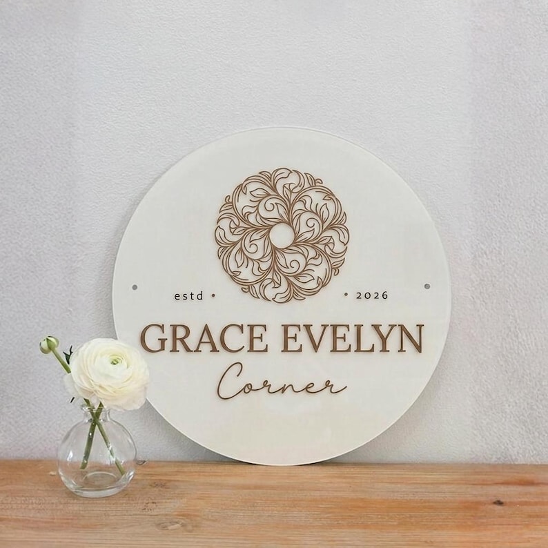 May include: A circular white sign featuring a brown floral mandala design at the top, the text "estd 2026" in the center, and the words "GRACE EVELYN Corner" printed below in brown. A small clear glass vase holds a single white ranunculus flower.