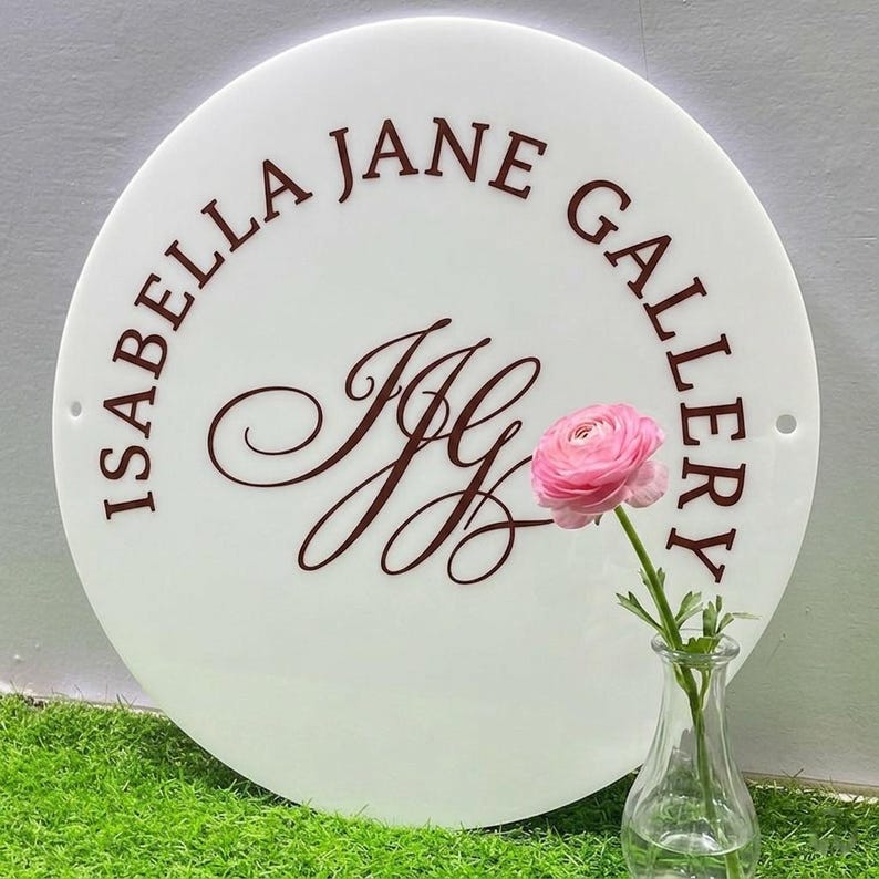 May include: A circular white acrylic sign featuring the dark brown text ISABELLA JANE GALLERY and a central monogram of the letters IJG in a cursive script. The sign has two small circular holes on the left and right sides for mounting purposes.