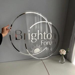 May include: A circular transparent acrylic sign featuring the silver text Brighton Forum and a stylized sun icon. The sign is framed by two overlapping silver circular outlines, creating a layered effect with reflective metallic lettering.