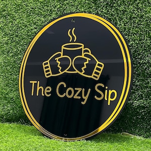 May include: A circular black sign with a gold border featuring a graphic of two hands wearing mittens holding a steaming mug. Below the graphic, the text "The Cozy Sip" is written in gold lettering. The sign has a small hole at the top.