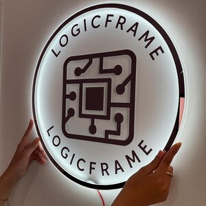 May include: A round, illuminated sign with the text "LOGICFRAME" encircling a central graphic of a circuit board. The sign is white with a dark border and the text and graphic are dark brown. The sign is being held by two hands.