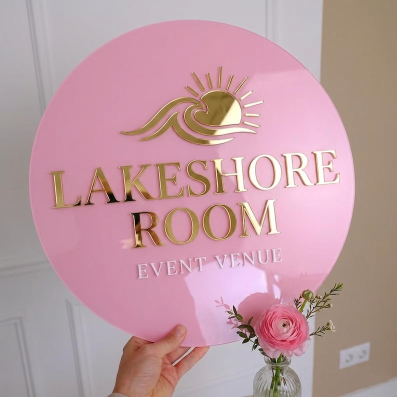 May include: A circular pink acrylic sign featuring a gold reflective sun and wave icon at the top. The text LAKESHORE ROOM is in gold, and the words EVENT VENUE are in white below. The sign has a smooth, glossy finish and reflective lettering.