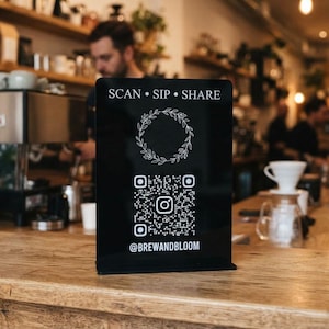 May include: A black rectangular acrylic sign with white text and graphics. The top reads SCAN • SIP • SHARE, followed by a circular leaf wreath graphic, a square QR code with an Instagram icon in the center, and the handle @BREWANDBLOOM at the bottom.