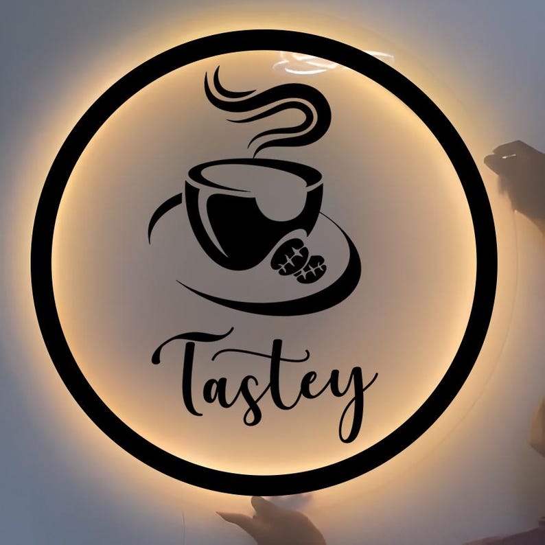 May include: A circular wall sign with a black frame and warm backlighting. The center features a black graphic of a steaming coffee cup on a saucer with two coffee beans. Below the graphic, the word Tastey is written in black script.