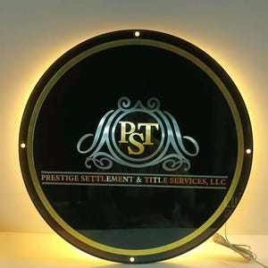 May include: A round, illuminated sign with a black background and gold trim. The sign features the text "PST" in a silver and gold design, with the words "PRESTIGE SETTLEMENT & TITLE SERVICES, LLC" below. The sign is lit with a warm, yellow glow.