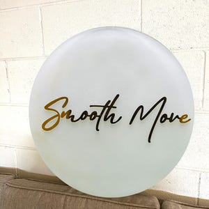May include: White round sign with the text "Smooth Move" in black script lettering with a gold outline.