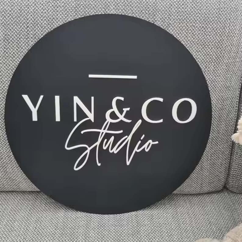 May include: A round, black sign with the white text "YIN & CO Studio". The text is arranged in two lines, with "YIN & CO" in a sans-serif font and "Studio" in a cursive script. A white horizontal line is above the "YIN & CO" text.