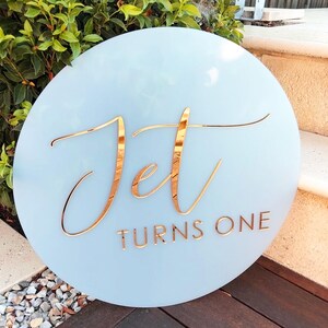 以下が含まれることがあります： 反射する金色の文字が施された円形の水色の看板。Jetという単語は大きな筆記体で書かれており、その下にTURNS ONEという単語がシンプルで清潔感のある大文字のサンセリフフォントで印刷されています。