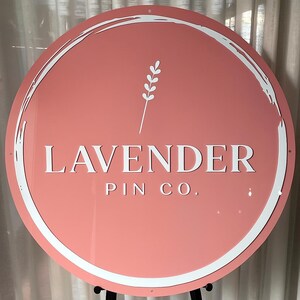May include: A round sign with a coral background and white text that reads "LAVENDER PIN CO." A white, stylized lavender sprig is above the text. The sign has a white, brushstroke-style border.