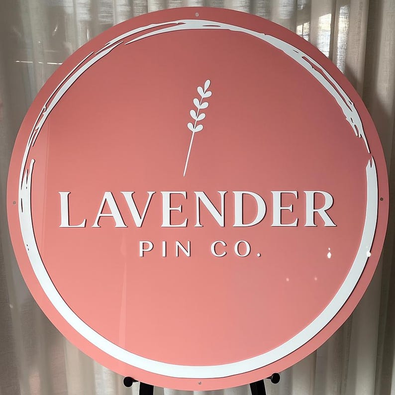 May include: A round sign with a coral background and white text that reads "LAVENDER PIN CO." A white, stylized lavender sprig is above the text. The sign has a white, brushstroke-style border.