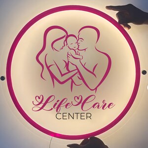 May include: A circular, illuminated sign with a pink border featuring a stylized graphic of two adults holding a baby. Below the graphic, the text reads Life Care in a cursive font, with the word CENTER printed in a simple, sans-serif typeface.