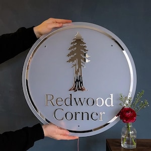 May include: A circular sign with a frosted finish featuring a metallic silver redwood tree graphic and the text Redwood Corner in a serif font. The sign has a polished silver border and includes small mounting holes at the top and bottom.