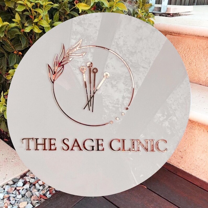 May include: A white round sign with a rose gold logo. The logo features a stylized branch with leaves and five needles. The text "THE SAGE CLINIC" is in rose gold below the logo.