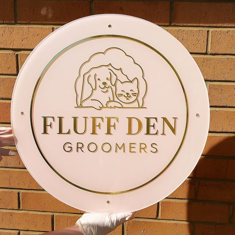 May include: A round, pink sign with gold lettering and accents. The sign features a gold illustration of a dog and cat, with the words "FLUFF DEN GROOMERS" in gold. The sign is held up by a gloved hand.