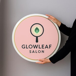 May include: A round sign with a pink background and gold trim, featuring a leaf design within a magnifying glass. The sign reads "GLOWLEAF SALON" in black text. The sign is held by a person.