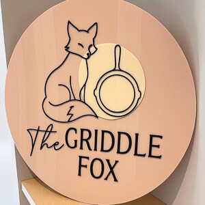 May include: A round sign with a light peach background features a black outline of a fox and a frying pan. The text "The GRIDDLE FOX" is in black script. The fox is sitting, and the pan is in a cream-colored circle.