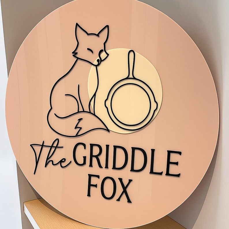 May include: A round sign with a light peach background features a black outline of a fox and a frying pan. The text "The GRIDDLE FOX" is in black script. The fox is sitting, and the pan is in a cream-colored circle.