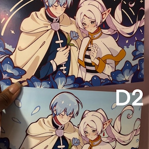 May include: Two anime-style prints, labelled D1 and D2, depicting a couple. The male figure has light blue hair and a cream-coloured cape. The female figure has white hair and a striped collar. Both are surrounded by blue floral elements.