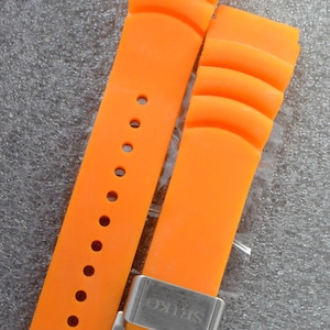 May include: Vibrant orange watch strap with a ridged pattern. The strap features several adjustment holes and a silver buckle, engraved with "SEIKO". The watch strap is made of a flexible material.