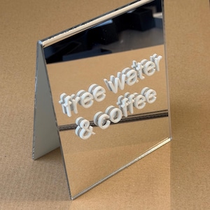 Op de afbeelding: Een spiegelend acrylbord met de woorden "free water & coffee" in witte, 3D-achtige letters. Het bord staat schuin en staat op een bruin oppervlak. Eenvoudig, modern ontwerp.