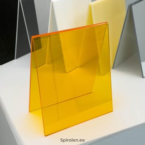 Könnte beinhalten: Eine Sammlung abgewinkelter Acryl-Displayständer in verschiedenen Farben, darunter Schwarz, Klar, Weiß, Gelb und ein leuchtendes Orange. Die Ständer sind auf einer weißen Oberfläche angeordnet und zeigen ihre unterschiedlichen Farbtöne und Transparenz.