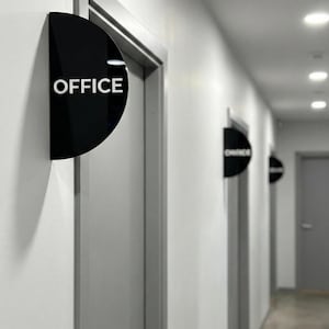 Könnte beinhalten: Ein Flur mit mehreren grauen Türen und schwarzen, halbkreisförmigen Schildern. Das erste Schild zeigt das Wort "OFFICE" in weißen Großbuchstaben. Die Schilder sind an der Wand neben den Türen angebracht. Die Wände sind weiß gestrichen.