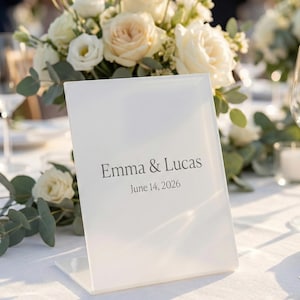 Op de afbeelding: Een witte tafelkaart met de namen "Emma & Lucas" en de datum "14 juni 2026" in grijs. De kaart staat op een wit tafelkleed, met een bloemstuk van witte rozen en groen in de achtergrond.