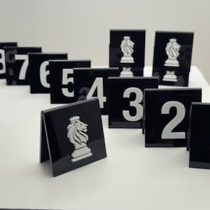 Op de afbeelding: Zwarte en witte schaakstukken. Elk stuk is een zwart vierkant met een wit nummer of een wit leeuwenembleem. De stukken zijn gerangschikt op een wit oppervlak. De nummers variëren van 2 tot 7.