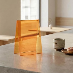 以下が含まれることがあります： オレンジ色のアクリル製卓上サイン。三角形に折りたたまれており、コンクリートの表面に立っています。サインは透明です。セラミックカップと食べ物の入った小さなプレートが背景にあります。