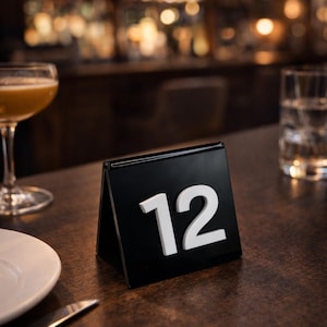 May include: A black table number stand displaying the number 12 in white. The stand is on a dark wooden table, with a blurred bar background. A cocktail and a glass of water are also on the table.