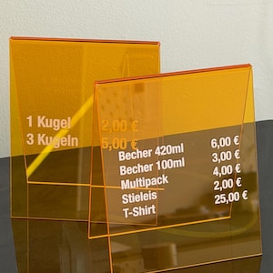 Könnte beinhalten: Zwei transparente orangefarbene Acrylschilder mit weißem Text. Die Schilder zeigen Preise für Artikel wie Eis, Getränke und ein T-Shirt. Die Schilder sind abgewinkelt, wahrscheinlich für ein Geschäft.