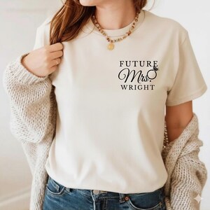 May include: A cream t-shirt featuring the text "FUTURE Mrs. WRIGHT" in black script, with a diamond ring graphic. The shirt is paired with a beige cardigan and blue jeans.