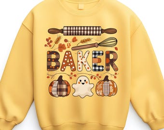 May include: Mustard yellow sweatshirt featuring a baking-themed design. The word "BAKER" is displayed with patterned letters. The design includes a rolling pin, whisk, pumpkins, leaves, and a ghost. A seasonal and festive garment.
