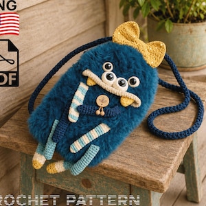 Puede incluir: Un bolso bandolera con forma de monstruo, de color azul oscuro y textura peluda, con un lazo de ganchillo amarillo, tres ojos negros y blancos, y brazos y piernas a rayas. Incluye una correa larga azul oscuro y una etiqueta redonda de madera.