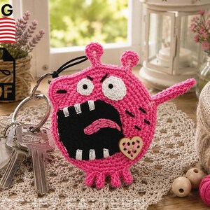 May include: A pink crocheted monster keychain featuring a wide black mouth with white teeth, a pink tongue, two white eyes, and a small wooden heart button. The monster has two small ears and three bottom feet, attached to a silver metal keyring with keys.