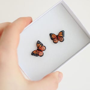May include: A pair of butterfly earrings in a white box. The butterflies are orange with black markings and are in a flying position.