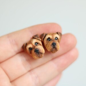 Puede incluir: Un par de pendientes de cabeza de perro de color marrón y negro hechos de arcilla polimérica. Los pendientes son pequeños y detallados, con rasgos realistas.
