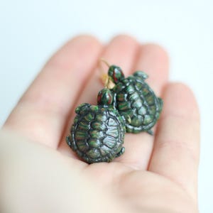 Puede incluir: Un par de pendientes con forma de tortuga. Los pendientes son de color verde oscuro con detalles rojos y dorados, y tienen un diseño de caparazón texturizado. Los pendientes están en un gancho dorado.