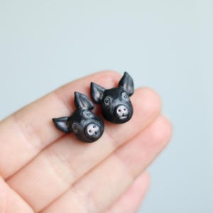 Puede incluir: Un par de pendientes de cerdo negro. Los hocicos de los cerdos están mirando hacia adelante y sus ojos están cerrados. Los pendientes están hechos de arcilla polimérica.