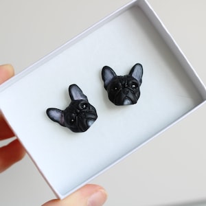 Puede incluir: Un par de pendientes negros con forma de cabeza de bulldog francés. Los pendientes están en una caja blanca, con la cara del perro detallada en negro y gris. Los pendientes son pequeños y parecen hechos a mano.