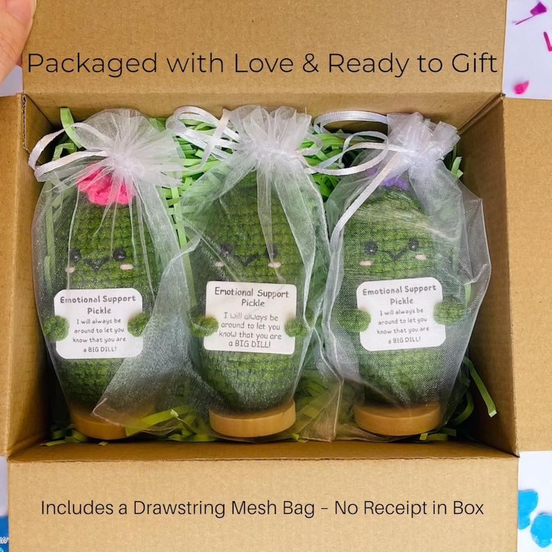 May include: Three green crocheted pickle dolls, each with a smiling face and a small tag that reads "Emotional Support Pickle." They are packaged in a cardboard box with green filler and a drawstring mesh bag. The text "Packaged with Love & Ready to Gift" is visible.