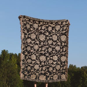 May include: A black and cream woven throw blanket with a floral pattern. The blanket is being held up by a person in a field.