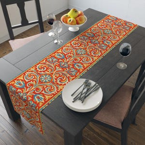May include: A rectangular table runner featuring a vibrant red background with a repeating pattern of swirling yellow and blue floral motifs. The runner is laid across a dark wooden table set with white plates, silverware, and wine glasses.