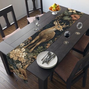 May include: A decorative table runner with a deer illustration, surrounded by floral and foliage patterns. The colour scheme includes brown, green, and beige tones. The runner is displayed on a dark wooden table, creating a sophisticated dining setting.
